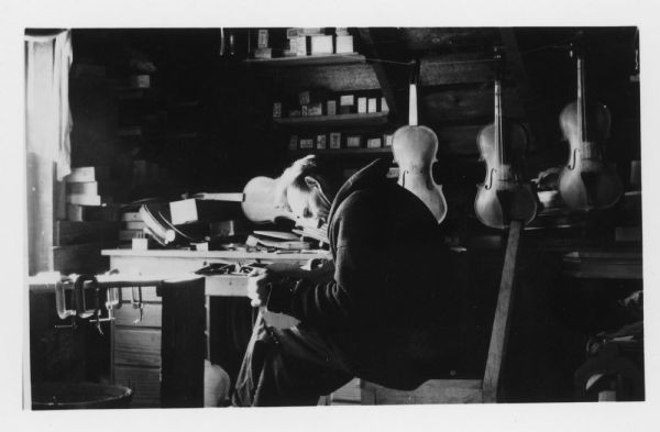 A man is sitting and hunching over the body of a violin. Other violins are hanging up behind him. Caption reads: "Louis J. Ropson, farmer & violin maker of Luxemburg, Wis. About 1940."