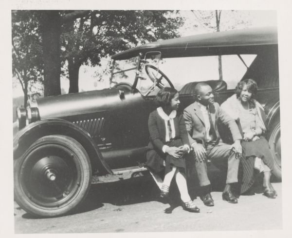 A man and two women sit on the running board of a car. Caption reads: "1st Car of Warren "Baby" Dodds, Negro drummer. 1923 Oldsmobile. Bright red. Price: $1470.40. L. to R.: Mrs. Tommy Ladnier [Daisy Matthews]; Tommy Ladnier, cornet player; Mrs. Warren Dodds (Irene Robinson). Louis Armstrong, noted trumpet player used this car to court his second wife, Lil. Copied from a photograph loaned by Warren Dodds, Chicago (via Larry Gara, Madison). N 2500."