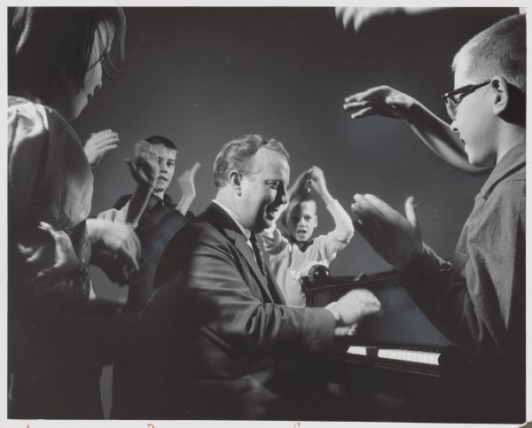 A man playing the piano while children around him are clapping their hands. Caption reads: "<b>Clapping out the beat,</b> children helped Tommy Sheridan, a Milwaukee pianist, in a jazz demonstration sponsored by the Children's Arts Program at the Memorial Center. Among the children were (from left) Kristin Liljequist, 7, of 1118 S. 37th st.; Terry Wendt, 11, of 910 E. Bradley rd., Fox Point; Curry Coffey, 8, of 2259 N. 64th st., Wauwatosa, and Eric Duhnke, 7, of 803 E. Otjen st."