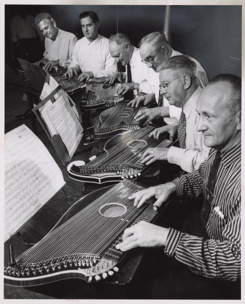 Six men playing zithers. Caption reads: "Members of the Alpenklaenge Zither club are a picture of concentration as they rehearse for their 20th spring concert at Jefferson hall today. They are, (from left): William May, Ludwig Muchlbacher, Randolph Mueller, Joseph Greiler, Albert Oberneder and Michael Buechl."