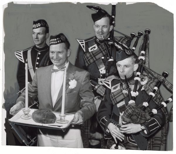 A man is carrying a tray with two candlesticks and haggis. Three other men are following wearing uniforms, and two men are playing bagpipes. Caption reads: "<b>Scots showed their own brand</b> of gemuetlichkeit — although they probably wouldn't call it that — at the annual Robert Burns dinner Saturday night at the Plankinton hotel. William Deuchars, 1755 S. Layton blvd., carried in the haggis. With him were members of the Rockford (Ill.) bagpipe band (from left) John Payne, Robert Stormont and Sigurd Aarli. The band played for the diners. Deuchars was chairman of the affair."