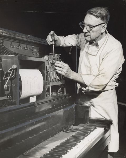 A man is using a screwdriver on a machine part of a player piano. Caption reads: "Rebuilding old player pianos keeps Elmer K. Hunholz busy now that the foot powered instruments are making a comeback."