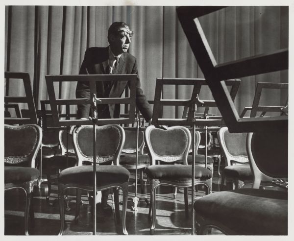 A man is leaning on the backs of two chairs, looking to the side. Caption reads: "Chairs — enough of them and in the right places — are a major concern for Harvey Ellerd, jr., 2171 E. Belleview pl., as stage manager for the Milwaukee Symphony orchestra. Ellerd also reigns backstage at productions of the Florentine opera company and the 'Music Under the Stars' concert series. Stage managing is his hobby."