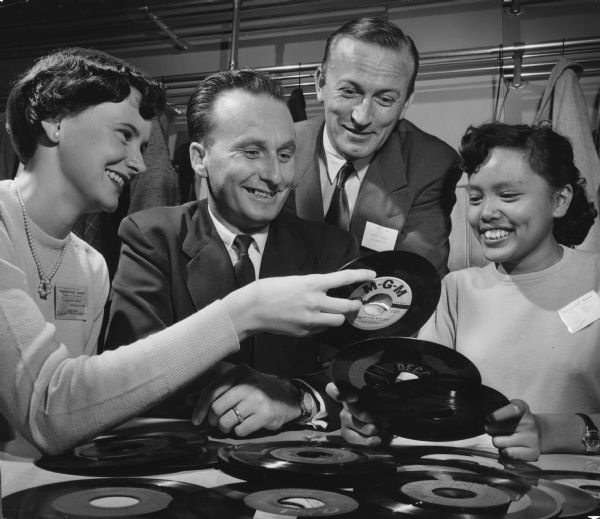 Two women and two men are looking at a pile of vinyl record albums. Caption reads: "<b>Foreign students</b> were among guests of honor at a mixer at Brooks Memorial Union, Marquette university, Thursday night. From left are Barbara McCarron, Medford, Wis., co-chairman of the mixer and member of the welcoming council; Steven Shiplov and Marko Pivac, Yugoslavian engineering students, and Mary Balmores, Honolulu, H.I."
