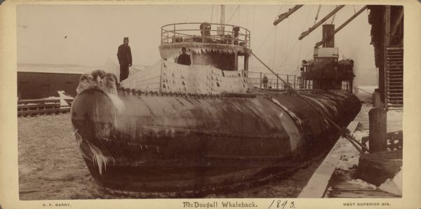 Men are standing in the foreground on the ice-covered whaleback docked in Superior. Signs on the pilot house at the back of the boat read "Mather" on the left, and "Samuel" on the right. Text at bottom right of card reads: "West Superior, Wis."