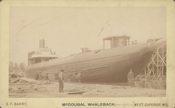 Text at bottom reads: "McDougal [sic] Whaleback. [manufactured at] West Superior, Wis." The steamer is under construction in the American Steel Barge Company shipyard in Superior, Wisconsin. Men are sitting and standing on the shoreline in the foreground.