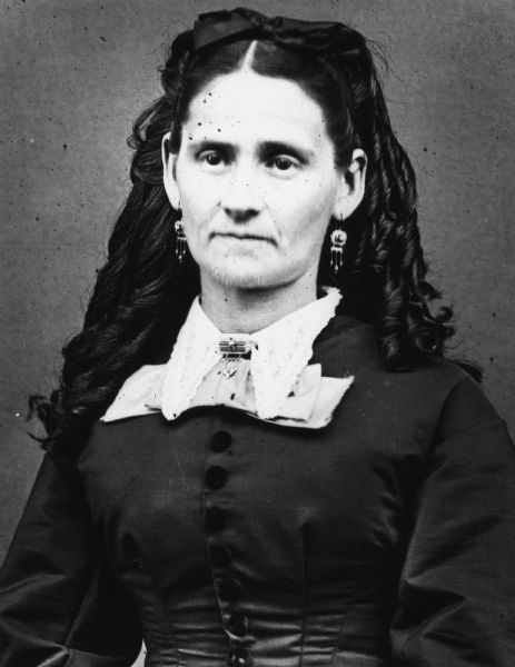 Seated portrait of a woman with curled hair, lace trim collar with pin, and earrings.