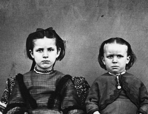 Detail of portrait of two unidentified children holding hands while posing on homemade fancy chairs. The girl is dressed in a hand-me-down dress cut from a heavy wool woman's dress, while the boy on the right is wearing an old-fashioned infant's dress which has become too small for him.