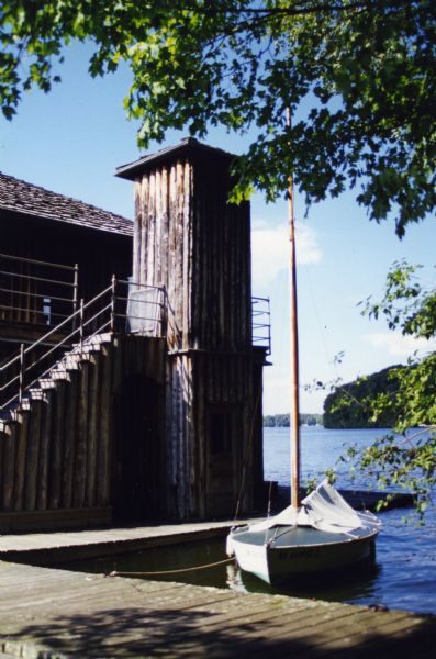 A vertical-log elevator shaft at one end of a vertical-log two-story boathouse; there is a door at the base of the elevator. Wood steps with a pipe railing lead to the second-story of the boathouse. A dock leads to and surrounds the boathouse that has a small sailboat moored to it. The boathouse is located on Lake Namakagon.