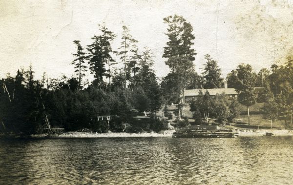 Cove Logging camp at future site of Forest Lodge property. The log building on the hill became the main house at Forest Lodge in 1902 when Crawford Livingston purchased the property. The hillside has a few very large pine treesm and smaller pines and birch trees. A log dock is at the water's edge.