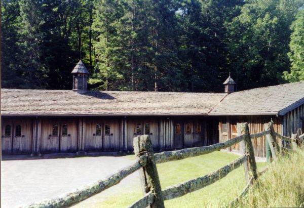 Single story wooden cow barn with an attached dairy to the right. Two cupolas are mounted on the cedar shake roof. A cedar rail fence separates the pasture from the driveway and barn. A forest of tall evergreen trees are in the background.