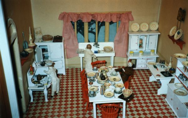 A fully furnished kitchen in a large dollhouse. The kitchen scene includes a doll chef at a stove with pots, pans and utensils; an icebox; a pedestal sink with a doll washing dishes; two side boards full of dishes; a fully set table with a doll diner; and a bench. The floor is a red and tan diamond pattern.