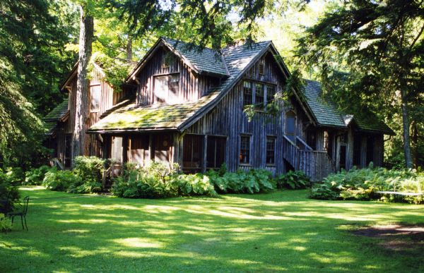 A side view over an expanse of mowed lawn toward the Forest Lodge guest house showing the screen porch and side entrance. The guest house is a two-story vertical log building that is surrounded by plantings and canopied by mature trees.