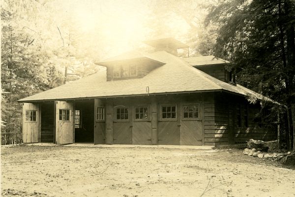 Four-stall two-story wood garage with four pairs of folding doors, each door with a six pane window; two doors are folded open. The roof has two dormers and a cupola. The barn is surrounded on three sides by large evergreen trees. The garage was destroyed by fire in the early 1920s. 