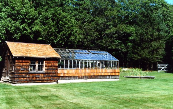 Cedar shake potting shed with an attached glass greenhouse located on a mowed lawn. A flower garden on the right is log-sided. Large trees surround the outer edge of the garden area.
