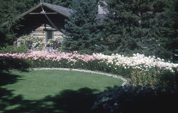 Circular flower garden with tall pink and white flowers as well as white alyssum edge a mowed lawn, near a main lodge entrance. Large hollowed log planters with pink flowers are located on the porch.