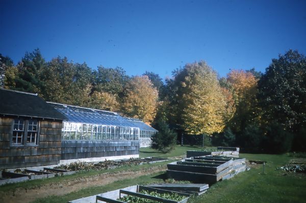 Glass greenhouse attached to a potting shed, with numerous cold frames on the right, all on a manicured lawn next to a deciduous forest.