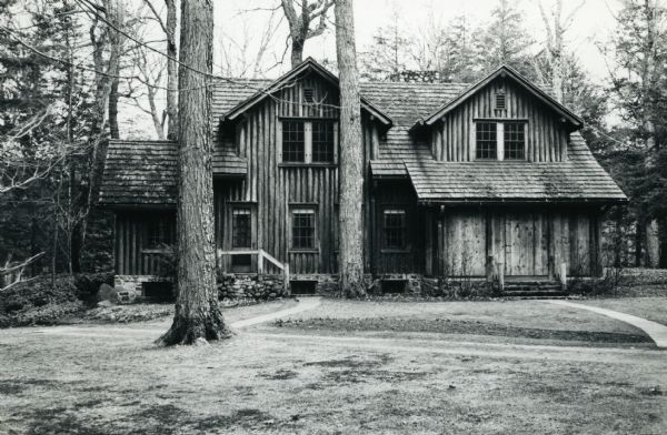 Two-story vertical log structure with cedar shake roof and a boarded over screen porch. The building is located in a woodland and lawn setting on the grounds of Forest Lodge.