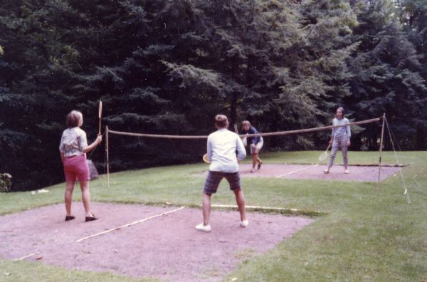 Two mixed-couples playing badminton on clay court at edge of evergreen forest. The two men and one woman are wearing Bermuda-style shorts.