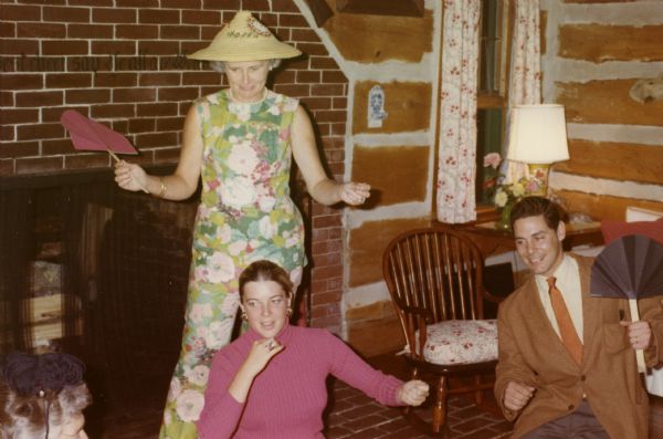 Two women and a man  are performing a skit in front of the brick fireplace in the living room at Forest Lodge. One woman is holding a paper fan and wearing a floral sleeveless pantsuit and a conical hat. The man is holding a paper fan and wearing a sport coat and tie.  