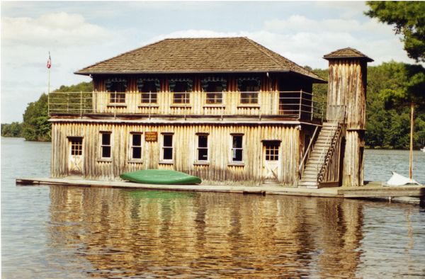 View of a two-story vertical-log structure surrounded by a lower deck and an upper deck with a pipe railing. There are ten double-hung windows, two wood doors, wooden stairs leading to the the second story, and a wood enclosed elevator shaft. There is a green canoe stored on the lower deck and a small sailboat docked at the deck. A flag is on a flagpole of the upper deck and there is a sign on the lower portion of the boathouse advising: "PRIVATE PLEASE KEEP OFF".