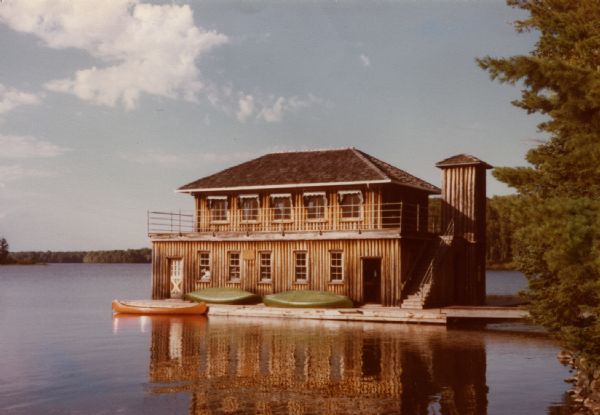 Two-story vertical log boathouse on Lake Namakagan. Two canoes are on the boathouse deck and a red canoe is in the lake tied to the deck. Both floors of the boathouse have windows, and the second story windows have awnings. A pipe railing surrounds the second floor deck. An elevator shaft is attached next to the outside stairs. A dock leads from the boathouse to the shoreline.