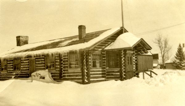 Single-story vertical log building with both a stone chimney and a brick chimney, as well as a front vestibule. Snow is on the roof and ground around the building.