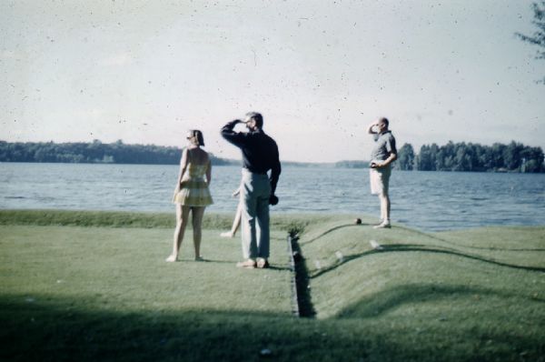Two couples standing at one end of the bowling green on the shore of  Namakagon Lake, searching down the field. One woman is wearing a yellow-checked skirted swimming suit. Two men are wearing casual summer attire. 
