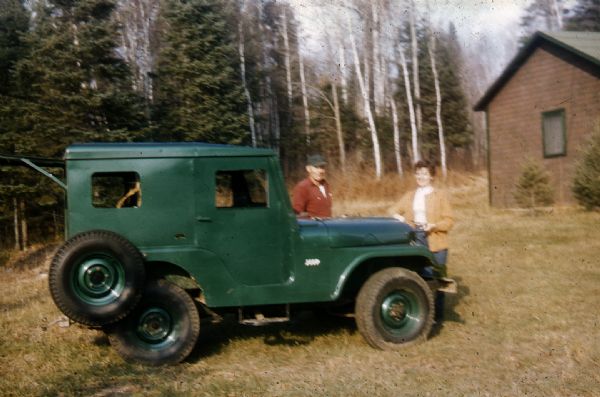 Man (probably Forest Lodge caretaker) and woman standing next to a green, enclosed Willys Jeep with a spare tire hanging on the passenger side. The man is wearing a red shirt and dark baseball-style cap. The woman is wearing slacks, white shirt and tan cardigan. The Jeep is parked in an opening of birch and evergreen trees, near a small one-story building.