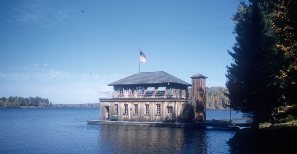 A two-story, vertical log boathouse on Lake Namakagon. The second floor has a wrap-around deck, and both floors have windows with green striped awnings. A vertical log outside elevator is attached to the boathouse, and a small sailboat is tied to the dock connecting the boathouse to the shore. An American flag is flying atop the boathouse.