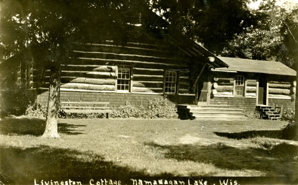 A horizontal chinked-log house with a wood porch and cedar shake foundation. A park bench is in the yard near the house which is situated among trees.