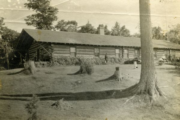 One-story long horizontal log building with one chimney. Four men are on a wood porch on the right, and one man is standing in the lawn holding a watering can. One large pine tree is standing among numerous stumps. A bucket with flowers is on top of one stump. There is foliage along the front of the cabin and trees behind the cabin. In 1902 this log cabin became the main house at Forest Lodge on Lake Namakagon.