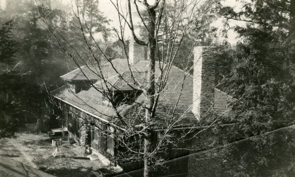 A vertical log house with cedar shake roof, two stone chimneys and two dormers. The house and front lawn are situated among trees.