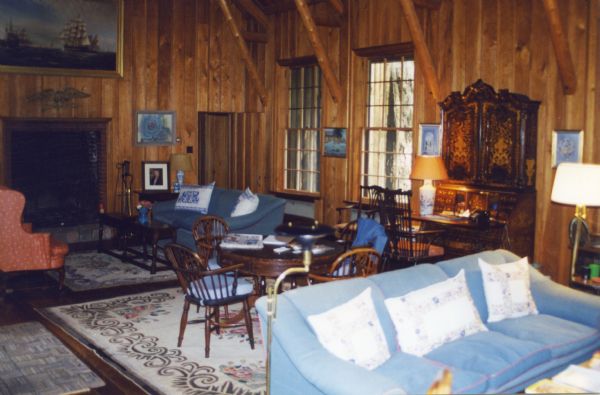 A large two-story wood paneled room with large casement windows and a fireplace that has a painting of a ship at sea hung above it. The  well-furnished room has two sofas, one a pale blue velvet with pillows, the other is a dark blue with pillows. An antique secretary desk is open with a push-button telephone on it. In between the two sofas is a round wood table and Windsor chairs sitting on a decorative wool rug. There is also an easy chair and antique coffee table arranged around a smaller wool rug. Numerous lamps are arranged around the room.