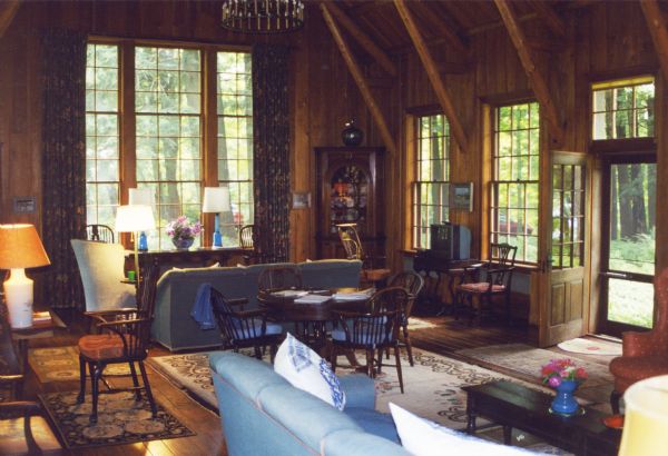 A wood paneled two-story room with log support beams, a chandelier, two large casement windows, a nearly two story triple sash window at the end of the room overlooking the forest, and an open front door with a transom. The room has two sofas, numerous lamps, and a round wood table and Windsor chairs on a decorative wool rug in the center of the room.