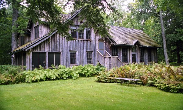 View of the two-story vertical log guest house with a screen porch, an outside stairway leading to the second story, and a variety of summer foliage surrounding the foundation. There is a metal and wood bench placed in front of a large planting of ferns in the mowed lawn.
