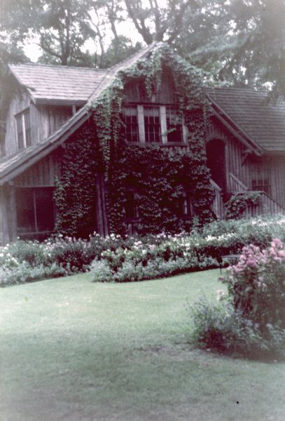 Two-story vertical log building with a screen porch, cedar shake roof, and a short flight of wood steps and railing leading to the entrance. Ivy is growing on the side of the building and long flower gardens are in the lawn.