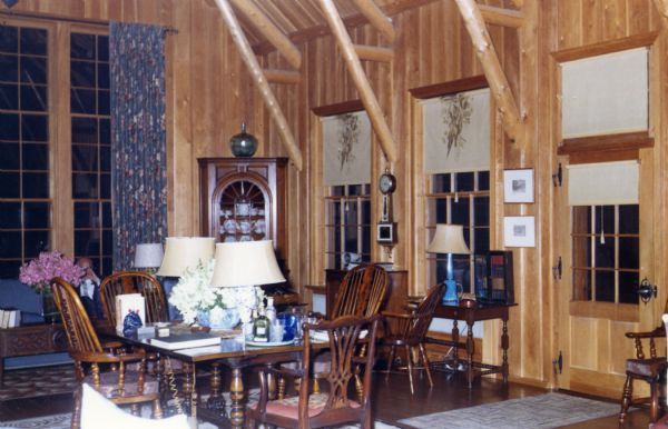 A light colored wood-sided, very tall room with wood support beams. A very tall window with curtains dominates an end wall, and the other wall has large sash windows and a door with roll-up shades. A wall clock is positioned between two sash windows.

A wood table and chairs is positioned across from the door and a smaller table is situated below one of the sash windows. Lamps sit on different tables and rugs are scattered on the floor. A corner hutch with pottery inside and a glass vase on top is in a corner next to the tall window.
  