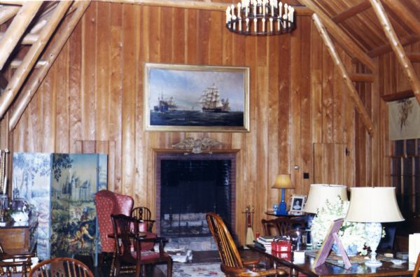 A tall, light-colored wood paneled room with exposed pine beams. A built-in fireplace with birch logs is on an end wall below a painting of ships at battle. A chandelier hangs above a wood table and chairs; lamps are on the table along with a bouquet of flowers and other items. A painted bi-fold screen is to the left of the fireplace.