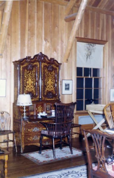An ornate secretary writing desk is positioned between two windows in the great room of the guest house. The desk is brown with gold inlays, two bottom drawers, a writing area and enclosed upper shelves. A Windsor dark wood chair sits on a rug that accompanies the desk. A lamp and a book or ledger sit on the writing section.