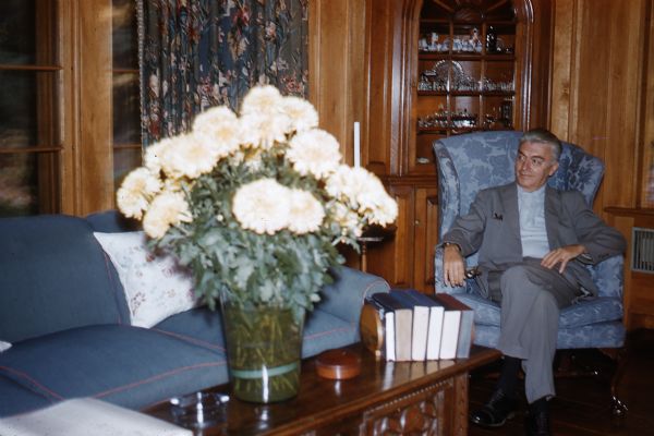 Middle aged man,possibly Jacskon BUrke, wearing a light gray suit with a light blue shirt, is seated, with his legs crossed, in a blue brocade upholstered wing-back chair in front of a built-in corner china cabinet with porcelain dishes located in the wood-paneled Great Room of the guest house. A vase of white chrysanthemums is on a table in front of a blue upholstered sofa.