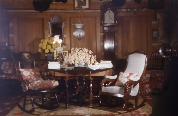 Chinked horizontal log-walled room with thick wool area rugs. Heavy dark wood rockers and Windsor chairs with seat cushions. A hanging light hangs over a Gateleg leg table. A grandfather clock is positioned against a wall between two doors, and two mounted bearskins are hung on the wall. Numerous pictures are on the walls.