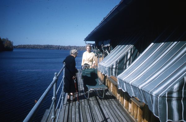 Mary Livingston Griggs, dressed in a black dress and coat and using a cane, is standing on the second story outside deck of the Forest Lodge Boathouse on Lake Namakagon, next to a green canvas and aluminum chaise-lounge. A younger blonde woman wearing a beige coat and dress is standing behind the chaise-lounge. The boathouse windows have blue-stripped canvas awnings; a pipe railing encircles the deck.