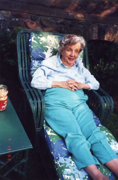 Mary Burke is lounging in a padded green wicker chaise lounge outside the main lodge at Forest Lodge on Lake Namakagon. There is a green wicker table with a jar of peanuts next to Mary who is wearing turquoise slacks and a print shirt.
On the back of the photograph is written: "Mary, here you are as we found you looking particularly pretty, feeding your chipmunk friends".