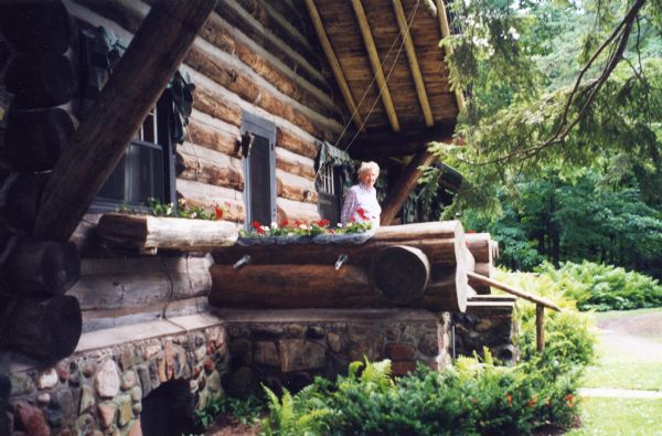 Mary Burke standing on the front porch of the main lodge at Forest Lodge on Lake Namakagon. The front porch is surrounded by ferns. Written on the back of the photograph: "As we say farewell, Mary makes warm the gate."

The main lodge is a horizontal log building with a stone foundation. The front porch is made of large logs that have flowering planters dug into them with spouts protruding out the sides for drainage. There are also flowering log planters under the lodge windows with cloth awnings.