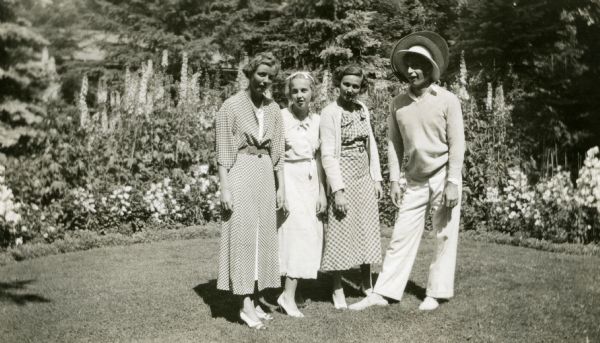 Three women, and one man wearing numerous hats, are standing on a lawn at Forest Lodge in front of a flower garden with woods in the background. The woman on the left is Mary Griggs (Burke) as a teenager.