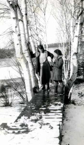 Mary Livingston Griggs and her daughter Mary are standing on a snowy birch-lined boardwalk on the shore of Lake Namakagon at Forest Lodge. Mother Mary is wearing boots and a wool coat and dress while leaning her right arm on a birch tree as she smilingly looks at the snow chunk her daughter is showing her. Daughter Mary is wearing boots, gators, coat, mittens and a hat as she holds up the snow chunk.