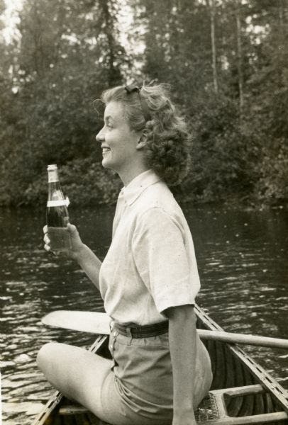 Mary is wearing belted shorts and a short sleeve blouse while sitting sideways in a wood-strip and canvas canoe that is at rest near the shoreline on Lake Namakagon. Mary is holding what appears to be a bottle of beer.
