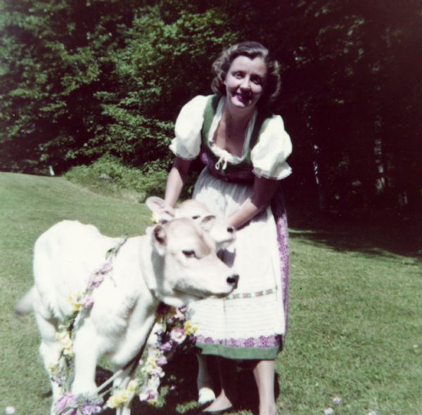 Mary Burke, dressed in a green dirndl with white short puffy sleeves and a white apron, is standing in the cow pasture at Forest Lodge holding two Brown Swiss calves wearing flower garlands.