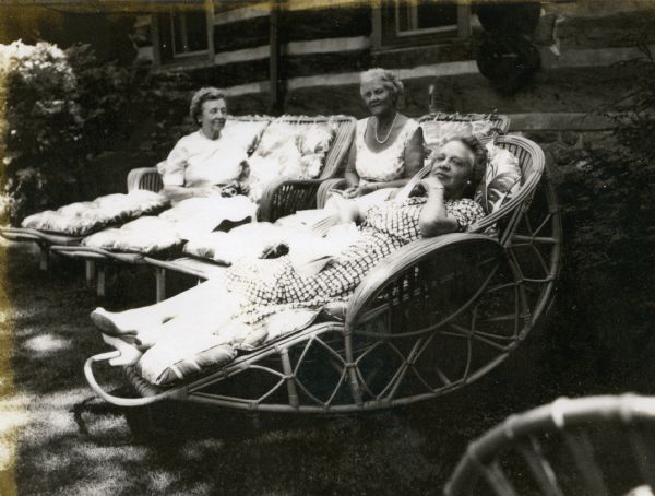 Mrs. T.W. Griggs lounging between two friends in wicker lawn chairs in front of Forest Lodge. One chair is a semi-circular rocking chair.